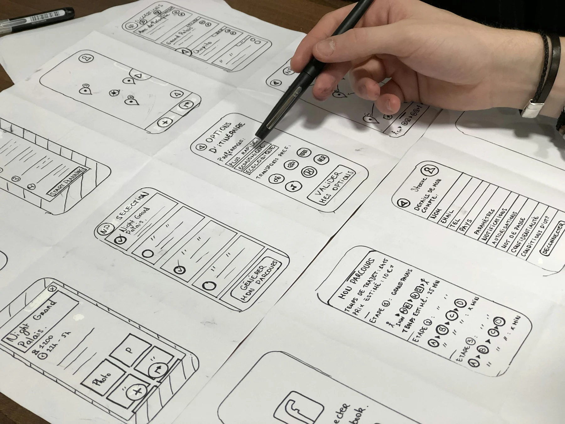 A hand points with a pen to hand-drawn mobile interface prototypes on paper.