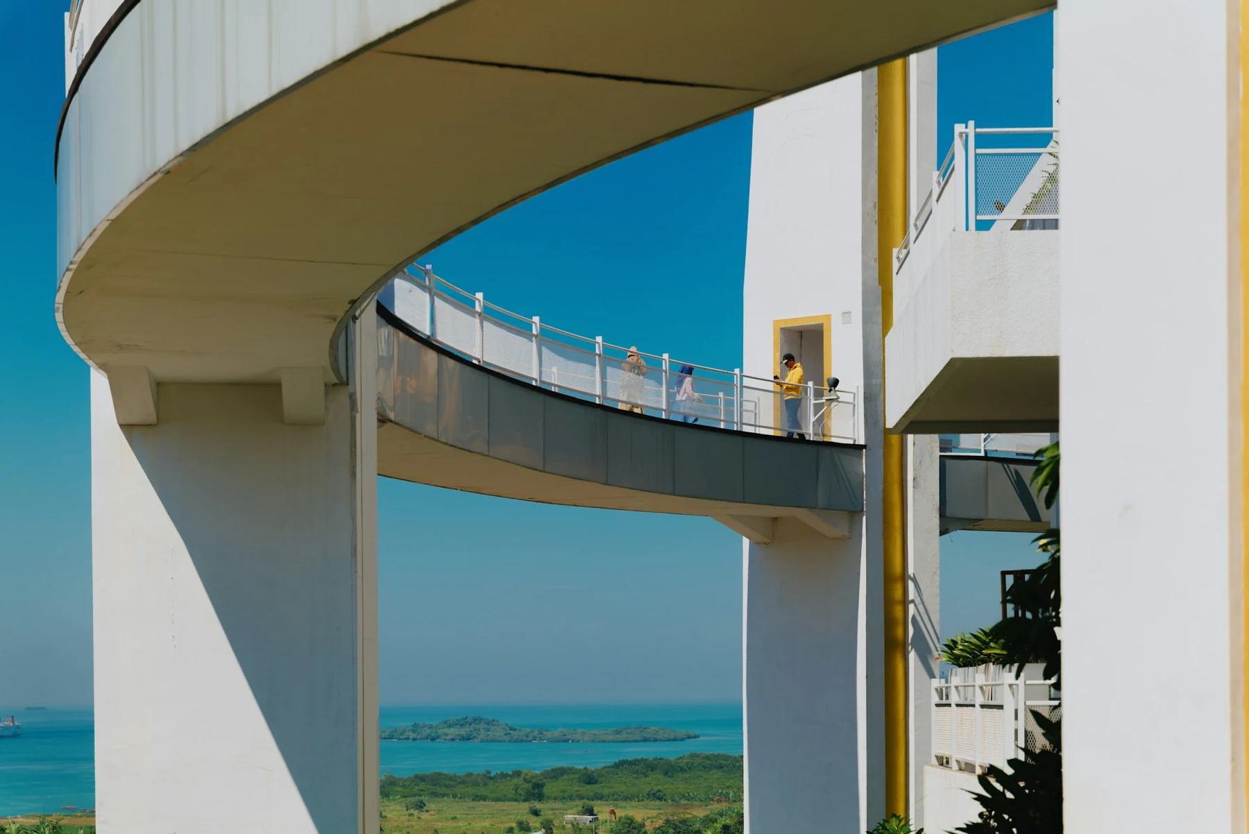 Curving white elevated walkways frame blue sky and a coastal view, with small groups of people moving through the architecture.